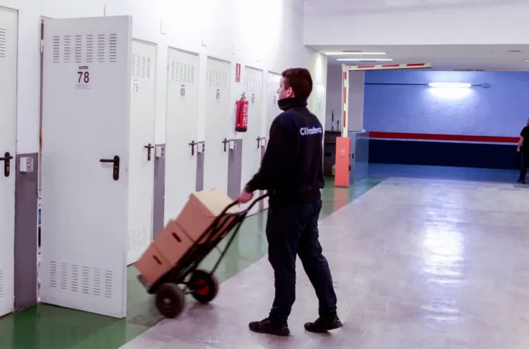 Una persona con uniforme oscuro empuja una carretilla cargada de cajas de cartón por un pasillo lleno de puertas blancas de trastero en el centro de Madrid.