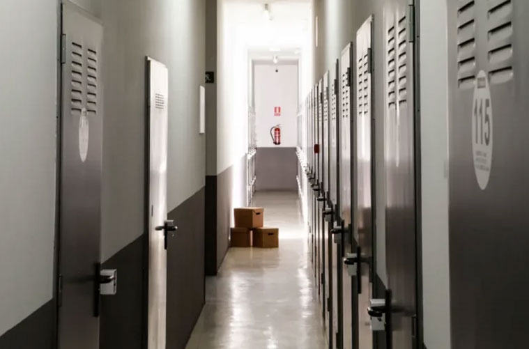 Un pasillo bien iluminado con puertas metálicas de trastero a ambos lados, cada puerta tiene rejillas de ventilación y números. Al final del pasillo en este trastero en el centro de Madrid, unas cuantas cajas de cartón se apilan cerca de una pared blanca y un extintor.