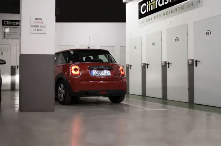 Un Mini Cooper rojo está aparcado en un trastero interior en el centro de Madrid cerca de unas puertas blancas rotuladas con números. La matrícula del coche es visible, y la zona parece limpia y bien iluminada.